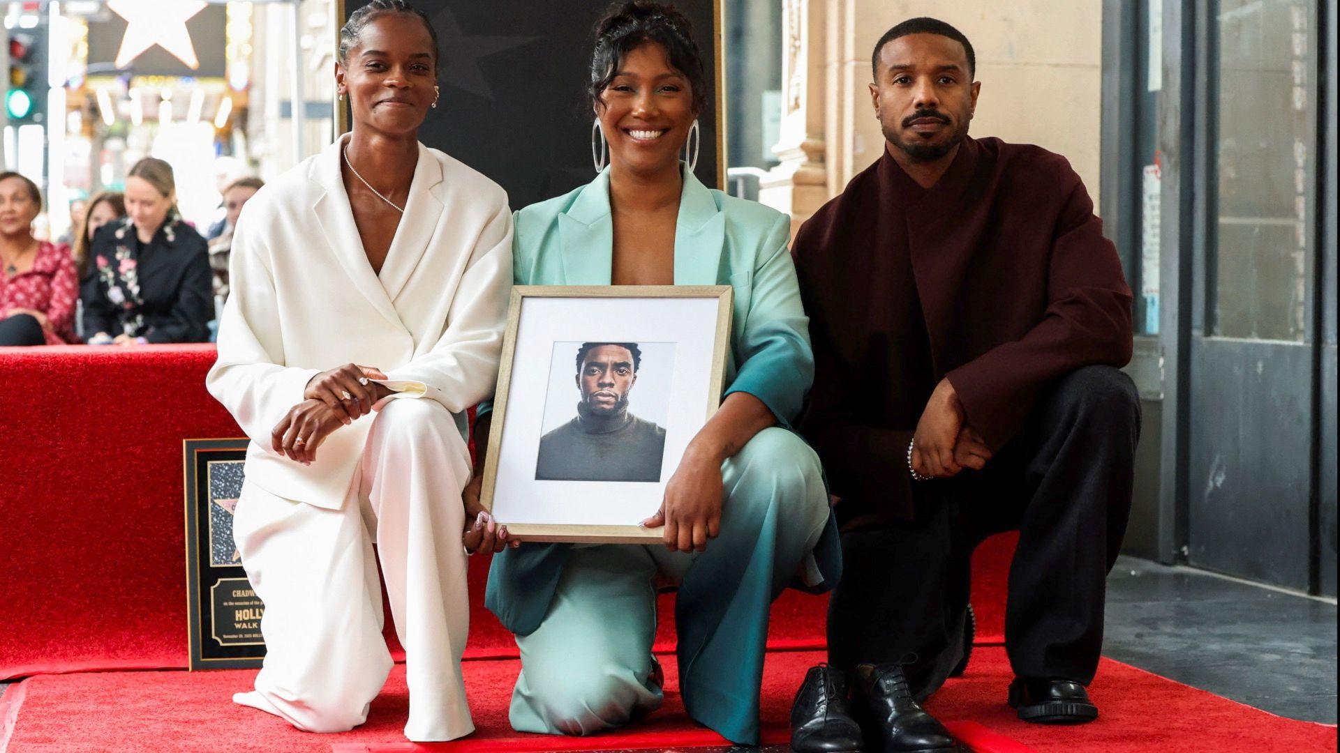 Three people kneeling smiling at the camera, two women are holding a photo of a man.