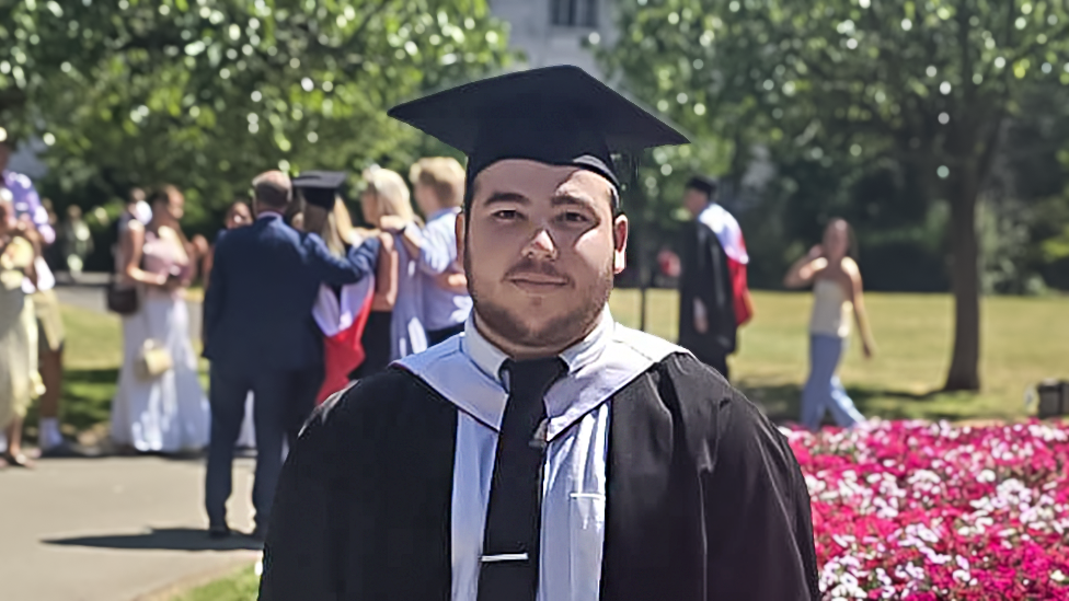 Oliver, who has dark hair and short facial hair, is wearing graduation robes and a cap, standing beside a flower bed in a park, with other graduates in the background.