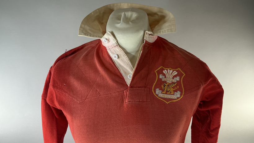 A Llanelli red long sleeved rugby jersey from the historic 1972 win over the All Blacks worn by Tommy David which is being sold at auction. It is shown on a mannequin bust with white background for auction catalogue. It has a white collar, with the club crest on the right hand side of the jersey.