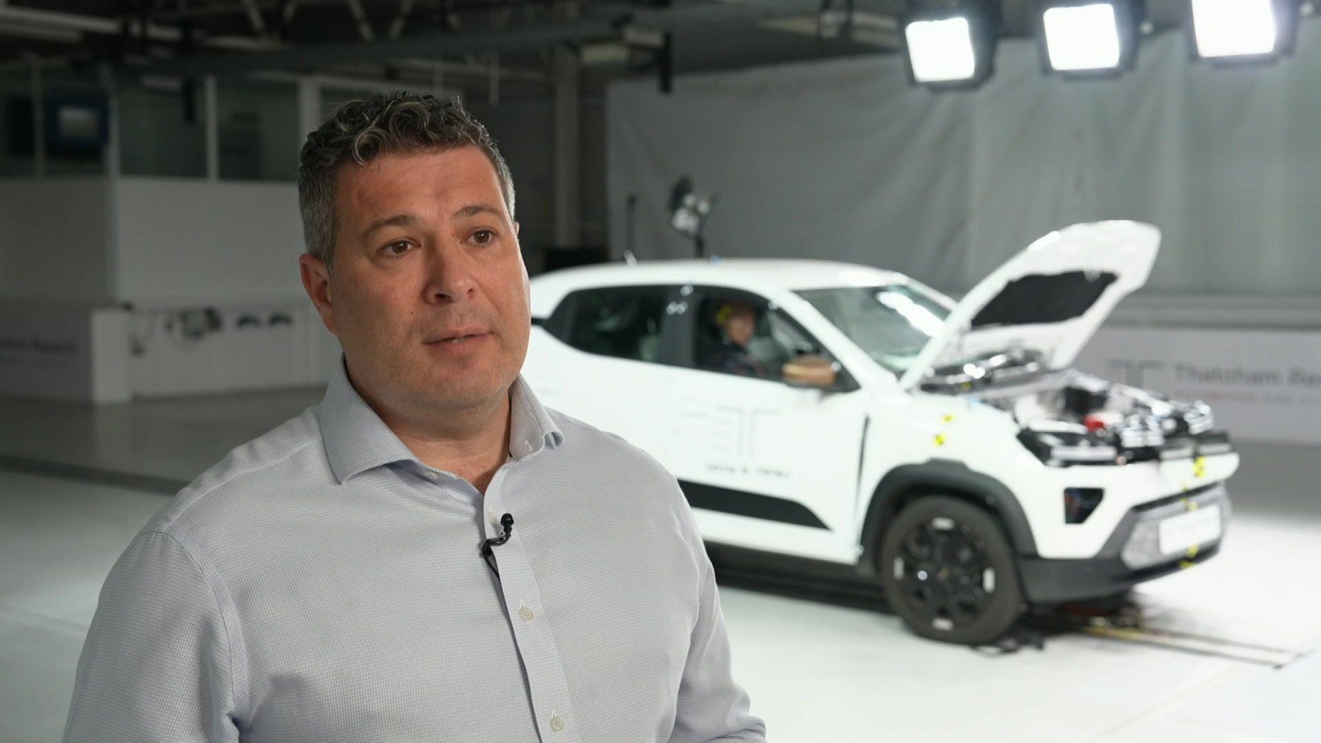 Thatcham Research's Dan Harrowell wearing a grey shirt with an open collar and no tie. Behind him in soft focus is the damaged EV mentioned earlier in the story with its bonnet lifted. He and the car are stood in the brightly lit Thatcham Research crash and safety testing laboratory.