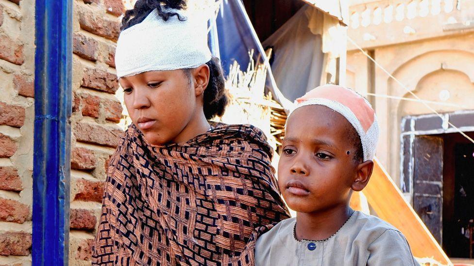 A woman and a boy injured during a drone strike in el-Obeid in North Kordofan sit outside a damaged property. Both of the heads are bandaged. They look solemn and stare away from the camera - 12 January 2026.