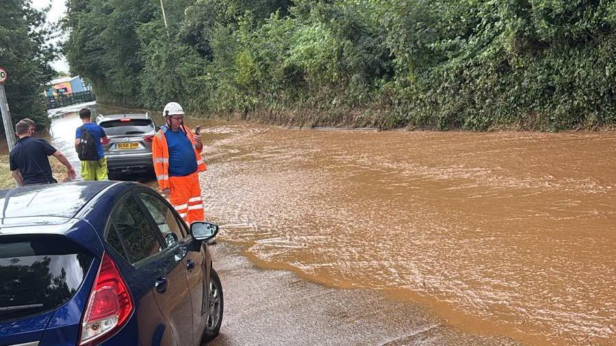 Flooding at Gwaelod-y-Garth, Taff's Well, Cardiff. Road full of mud and stones. Worker in orange hazard wear looking at the brown water.