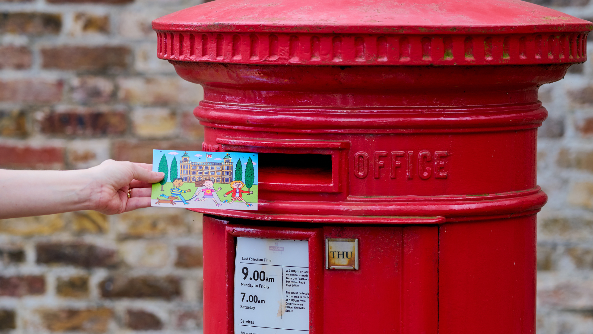 A hand holding a postcard with an illustration of a palace on it reaches towards a red postbox.