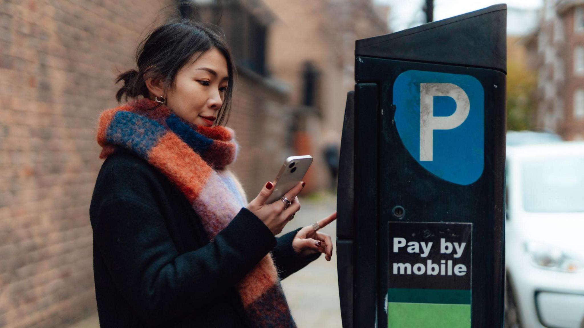 Young woman holding smartphone, making mobile payment at the parking payment machine.