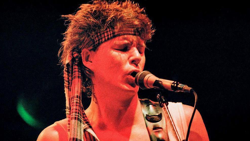 Stuart Adamson of Big Country singing into a microphone in a photo from the eighties. He has a spikey mullet and a tartan scarf tied round his head