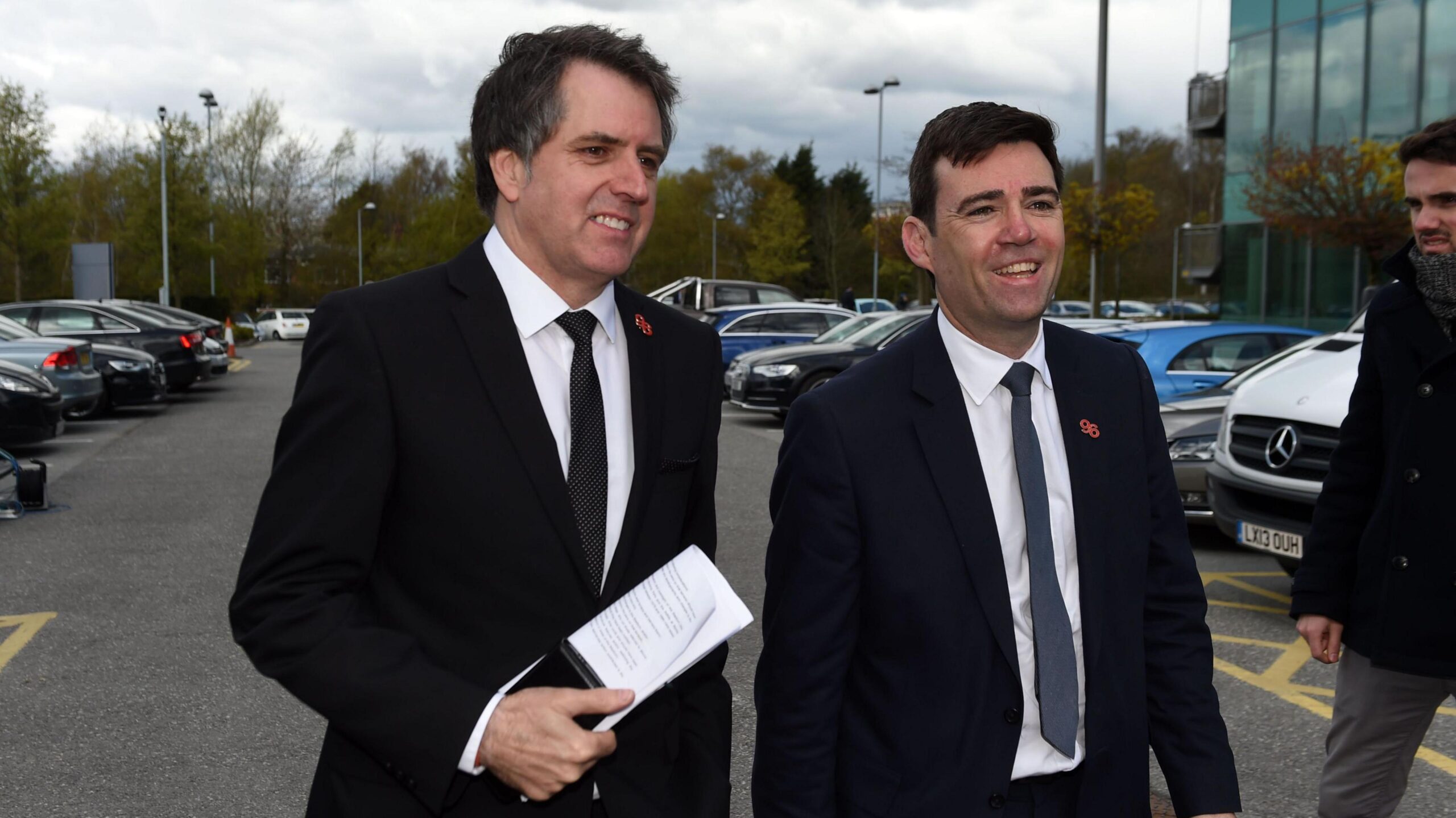Two white middle aged men with short hair wearing dark suits, white shirts and ties walk through a car park, smiling broadly.
