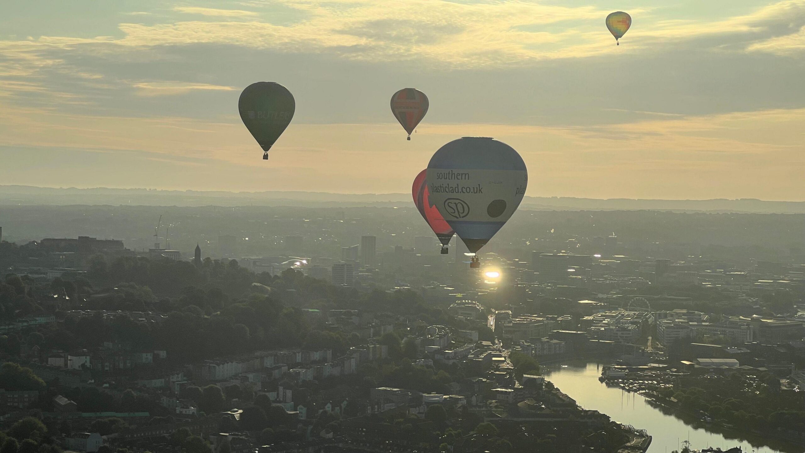 Hot air balloons flying over Bristol. The city and its harbour can be seen below, while the sun rises over the landscape.