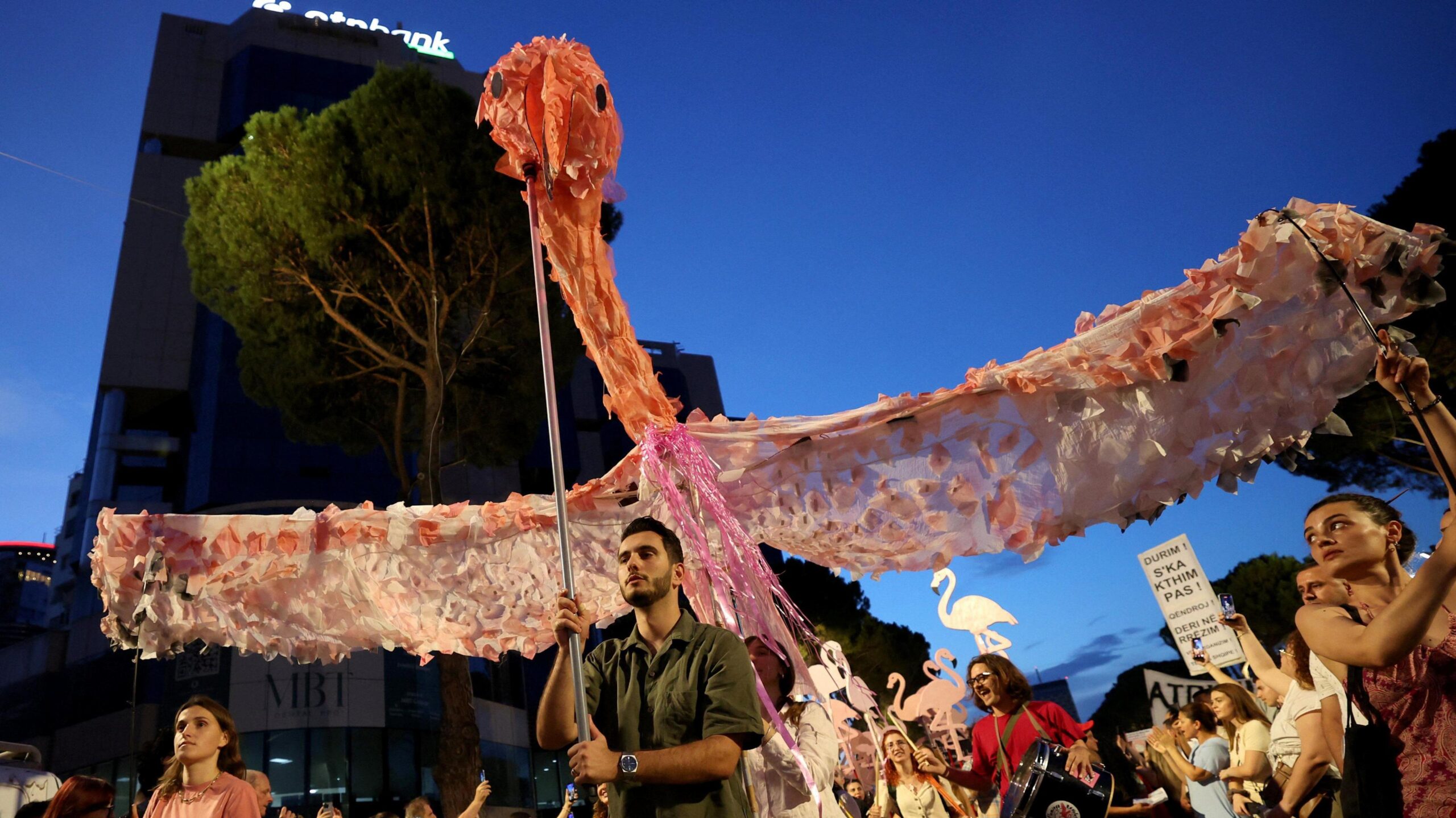 A man carries a large home-made puppet of a pink flamingo, flying above his head in a crowd of people protesting.
