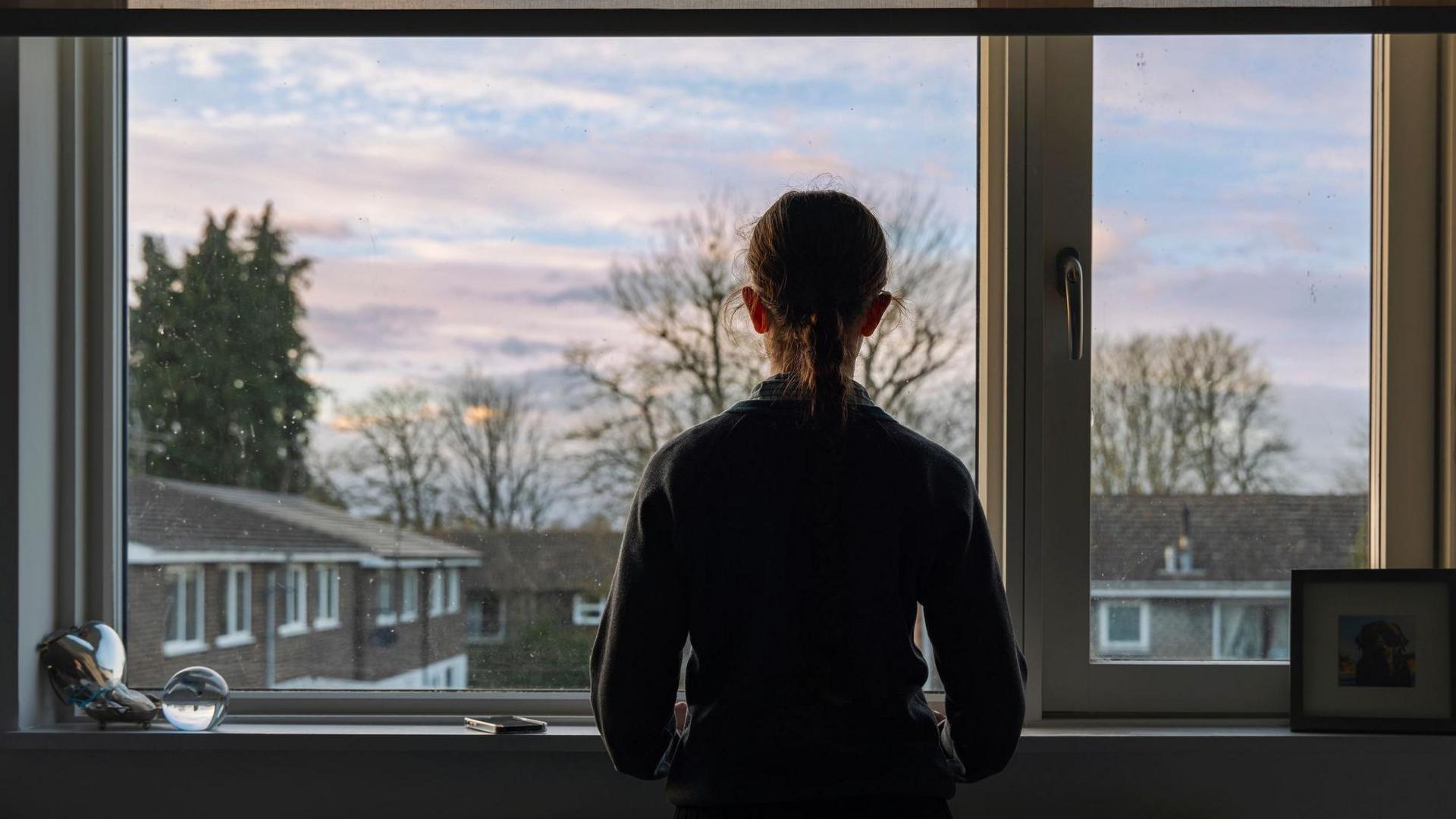 A young person with long hair tied in a ponytail looks of her bedroom window. They are in shadow, while the roofs of other houses are clearly visible and there are trees in the background.