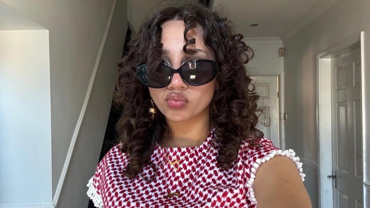 Mariam smiles at the camera and is wearing sunglasses with a white and red top. She has curly brown hair