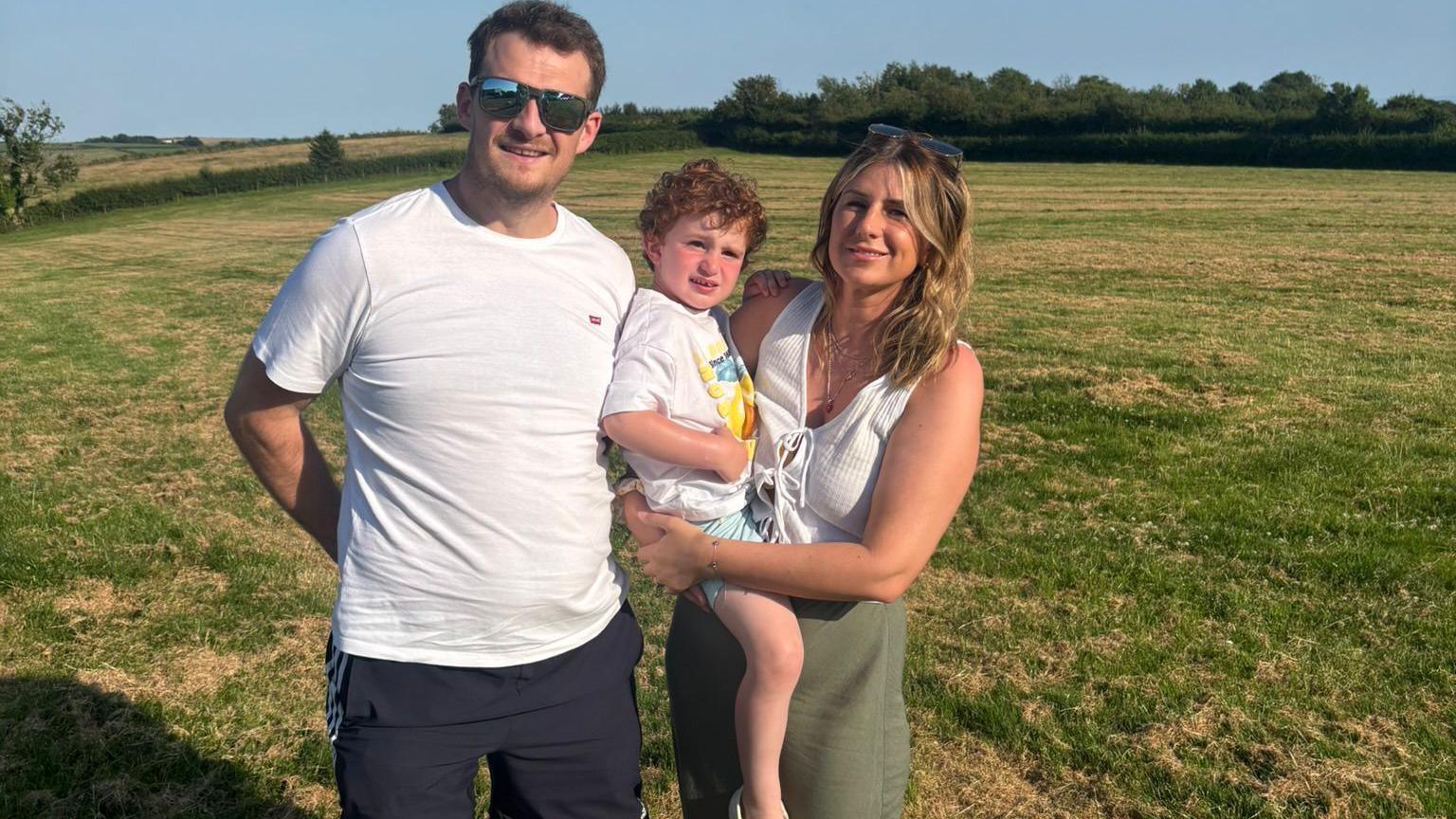 Richard stands with  his family in a field on a sunny day. He has short dark hair and reflective sunglasses and a white t-shirt. A woman with blonde hair, her sunglasses on her head smiles beside him. She is holding a boy on her hip, with Richard leaning into his back. The boy has curly red hair.
