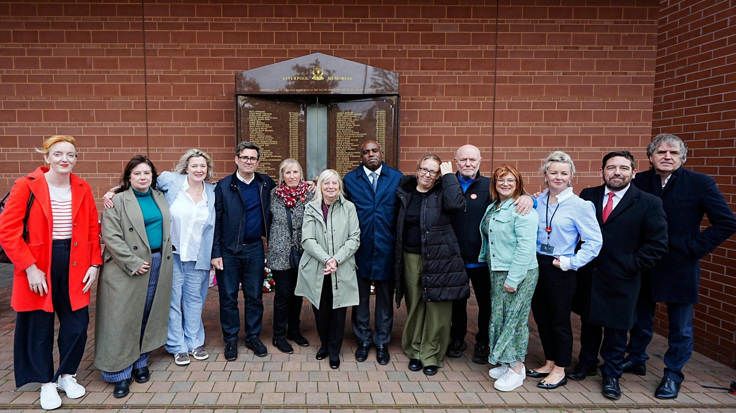 A group of 13 men and women stand in a line in front of a memorial plaque to the 97 Liverpool fans who were killed at the Hillsborough disaster in 1989.