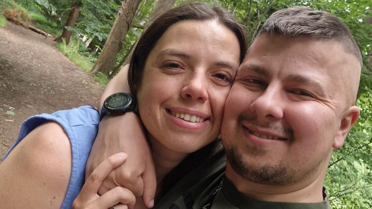 Josh, a man with brown hair and stubble wearing a green t shirt, taking a selfie with his arm around the neck of a brunette woman, Kelly, wearing a pale blue vest top and holding her hand over his. They are stood in a forest with trees in the background and are both smiling and looking at the camera.
