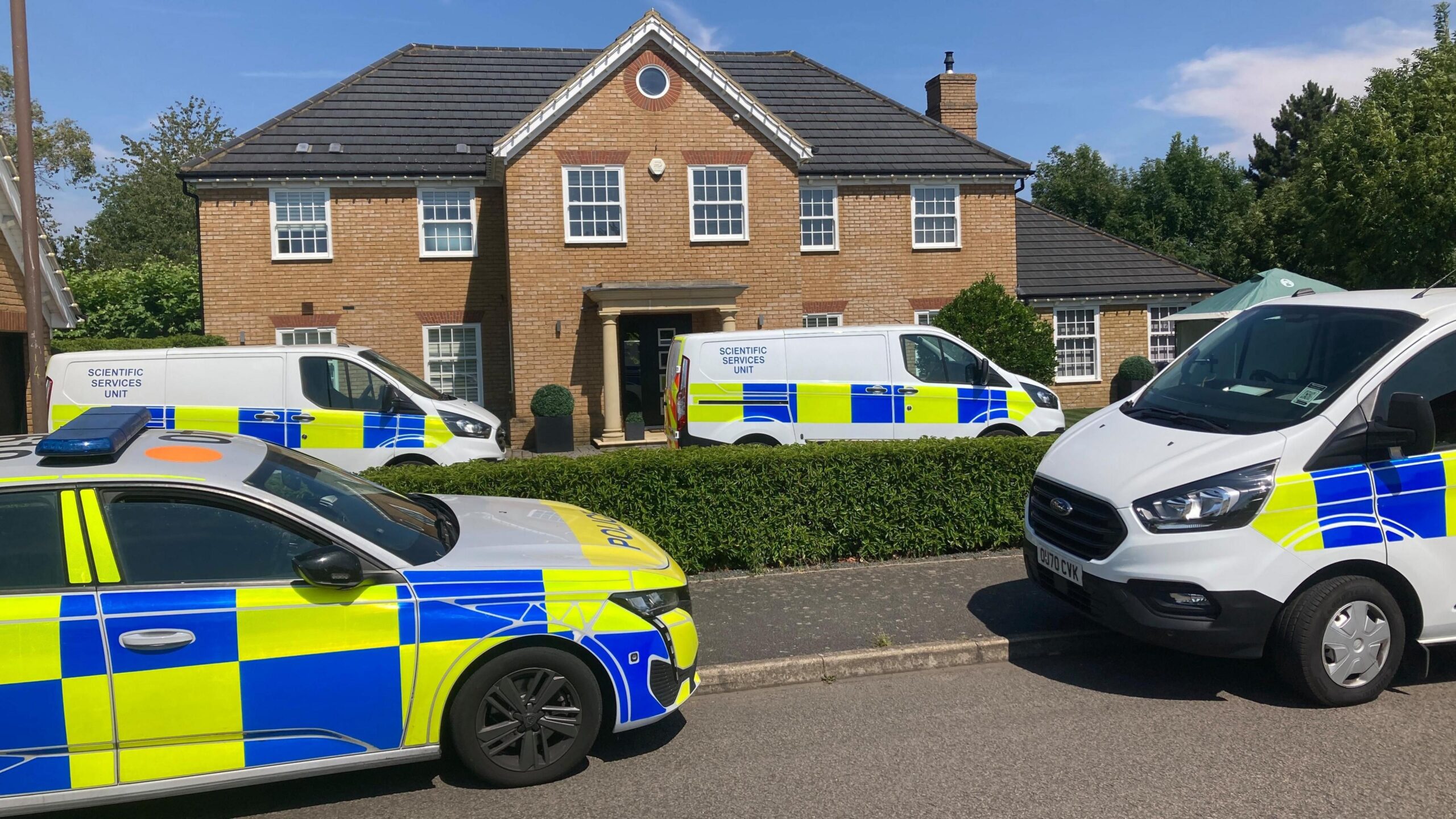A police car and a few forensic vans are parked outside a large house, some on the street, some on the driveway