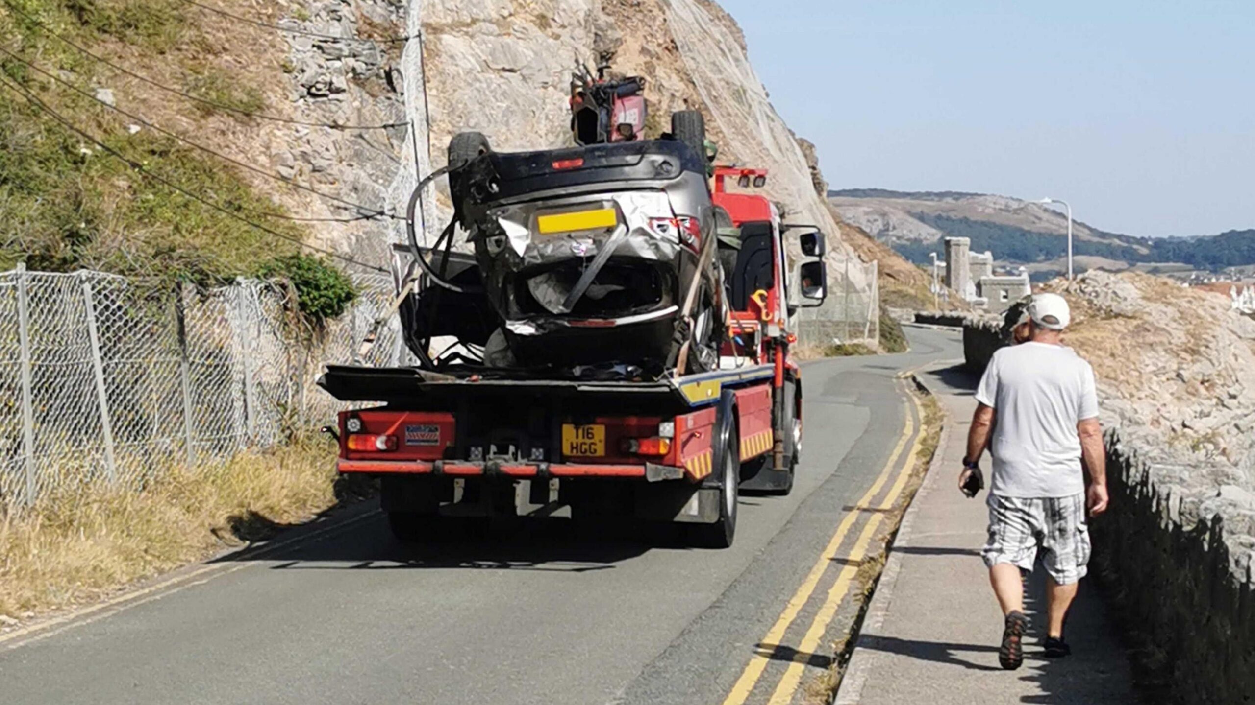 Upturned silver car fixed to back of red low loader and driven away as passersby look on