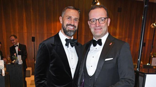 Jens Spahn (R) in glasses, a grey jacket and white shirt and bowtie beside his husband Daniel Funke who has as beard and a black velvet jacket with a white shirt and bowtie