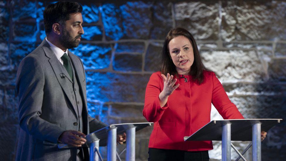 Humza Yousaf, who has dark hair and is wearing a grey suit, stands side on to Kate Forbes, who has shoulder-length dark hair and is wearing a red jacket. They are speaking at podiums in a colourfully lit television studio