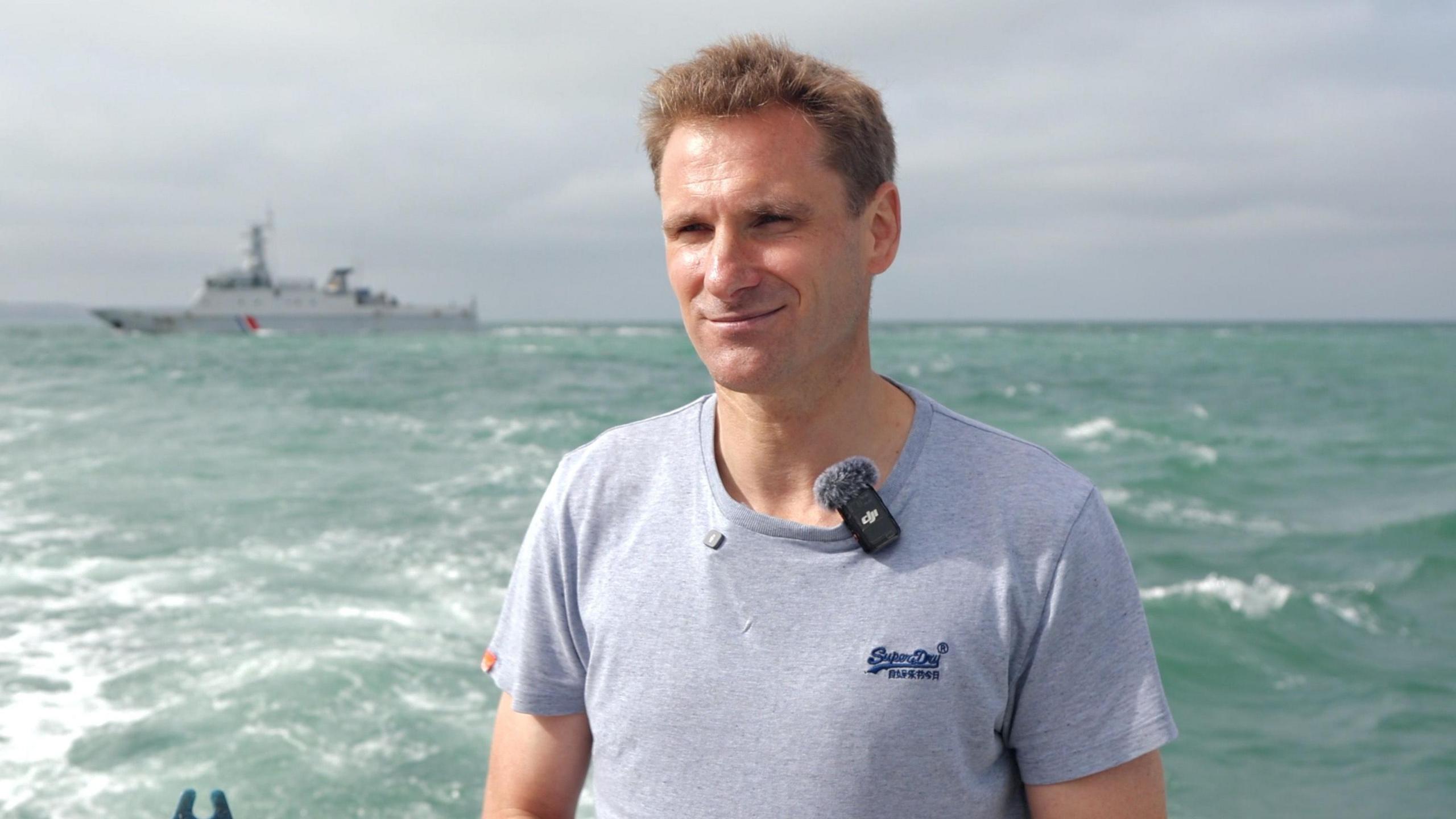 MP Chris Philp, in a grey t-shirt, stands on a boat in the sea. A large, grey ship is visible in the background.