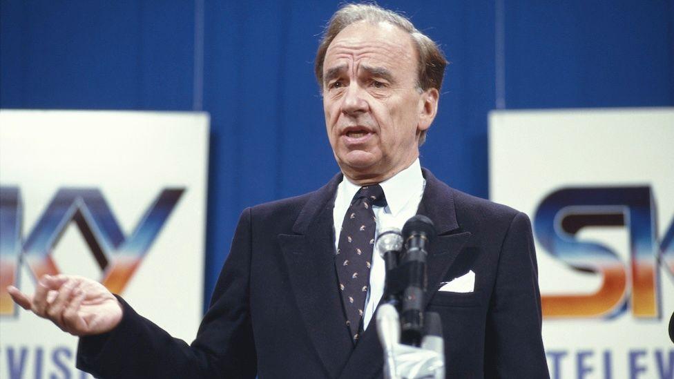 Rupert Murdoch pictured at the launch of Sky Television in 1989. He is dressed in a black suit and black tie with white flecks on it. The Sky logo is seen behind him.