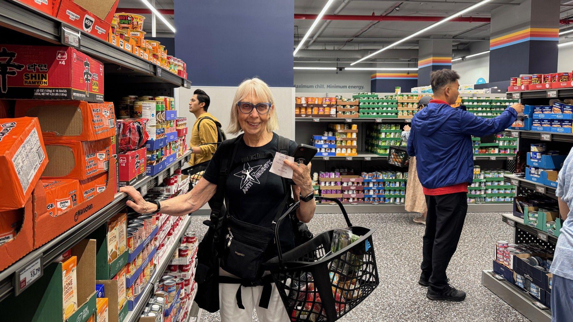 A woman with glasses in a black shirt and white pants holds a shopping basket