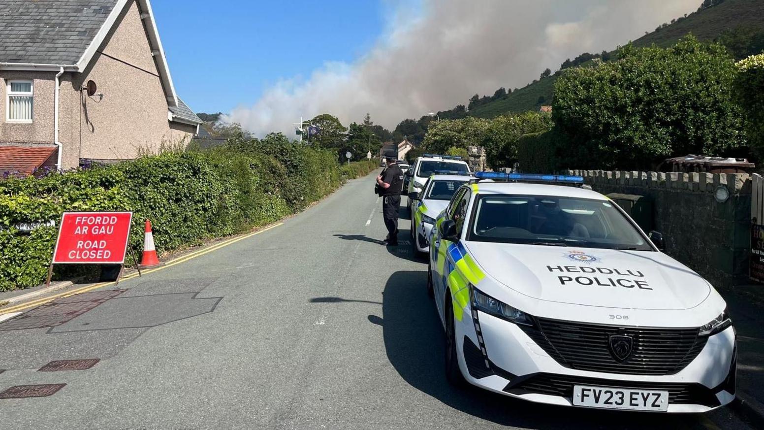 Police cars are parked on the right a road close sign can be seen on the left. There is a huge cloud of smoke rising into the sky over the hillside with houses seen on the street.