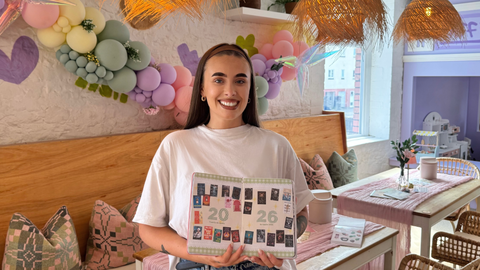 Amelia Lord holding a book or folder open with the numbers 2026 across it and images of books she has read or wants to read in that year. She has long straight hair held back by a headband, wears a white tshirt and jeans and is smiling at the camera. A bench with soft patterned cushions is behind her with two tables draped with soft pink cloth and vases of flowers. There is a balloon display on the walls in pastel colours.