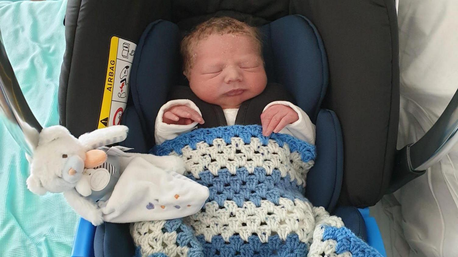 A baby wrapped in a blue and white blanket with a white teddy next to him