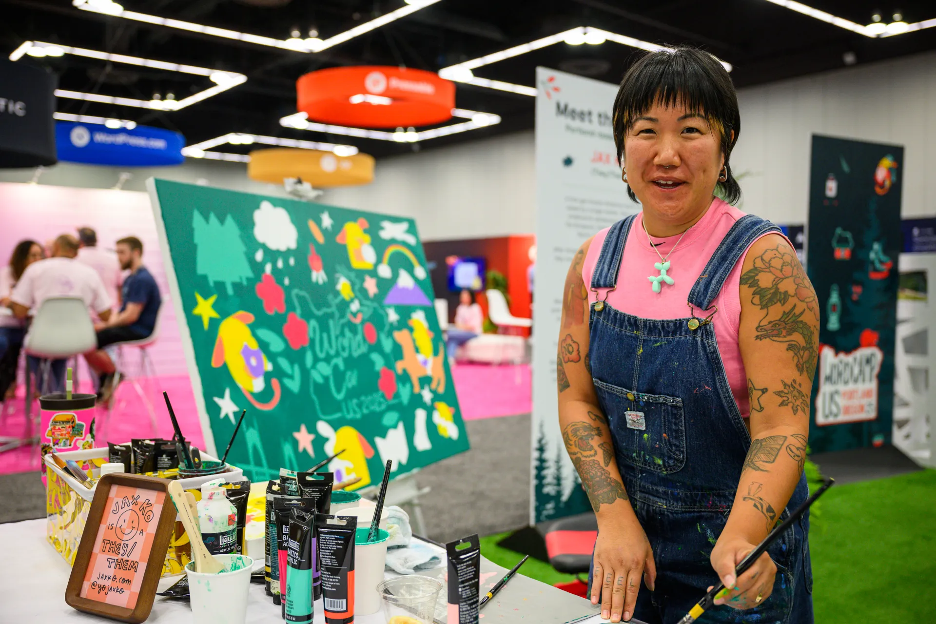 An artist stands beside a painted canvas at a WordCamp booth, holding a paintbrush.