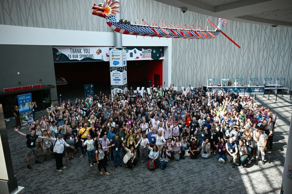 A large crowd of WordCamp US attendees waving for a group photo beneath a decorative dragon boat in the convention center lobby