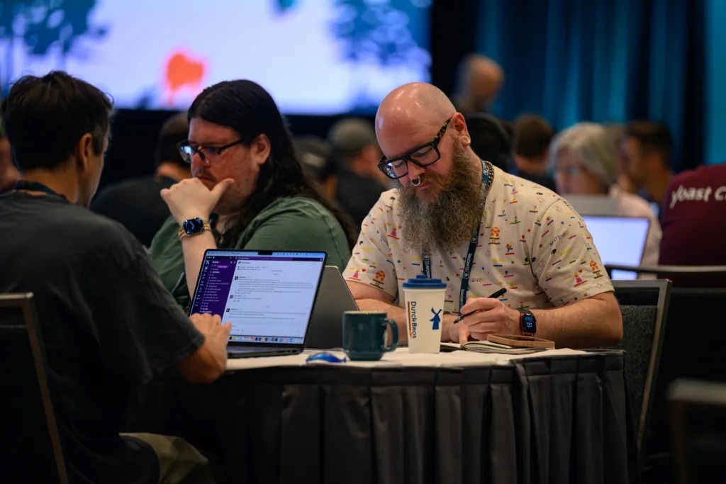 Three WordCamp US attendees working together at a table, one taking notes by hand while another types on a laptop