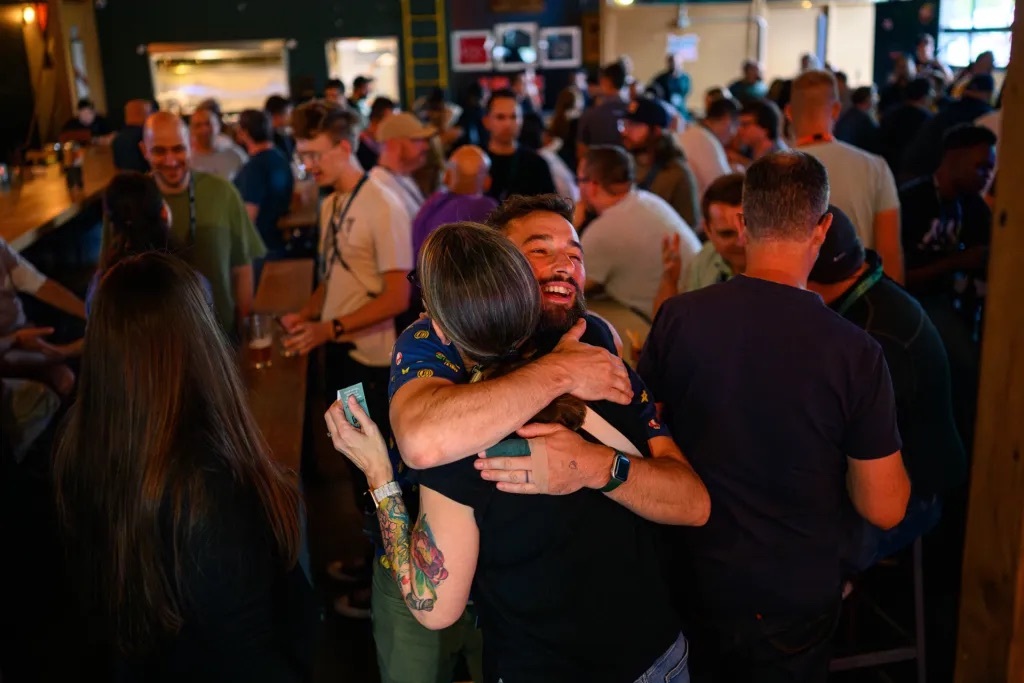 Two WordCamp US attendees hugging and laughing at a crowded evening social gathering
