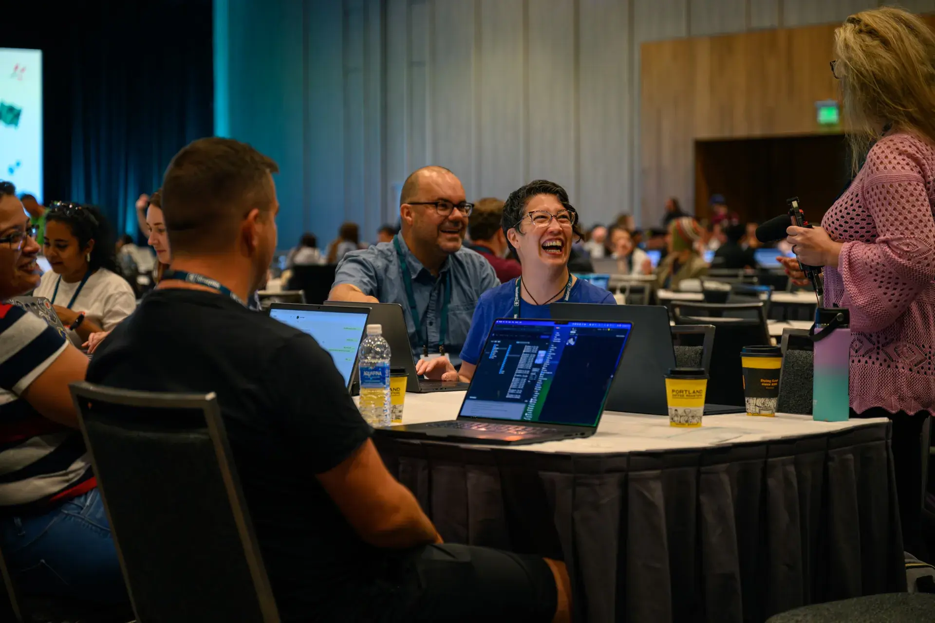 A group of WordCamp US attendees laughing together around a table of laptops and coffee cups while one person films with a microphone