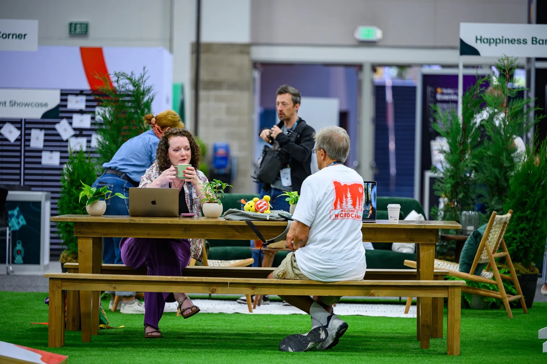 Two WordCamp US attendees chatting over coffee at a wooden picnic table on artificial grass near the Happiness Bar, with a Wapuu plush toy on the table