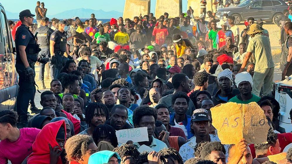 Migrants sat on a beach as police officers watch on, with some holding signs saying 'Asilo'