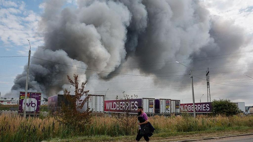 Wildberries employee walks past plumes of smoke rising above wildberries logistics  complex. There are seven company lorries in front of the smoke as well as roadside shrubs