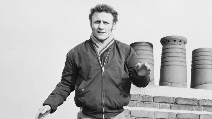 Paddy Maguire holds up his arm and stands in front of a row of chimneys in 1973