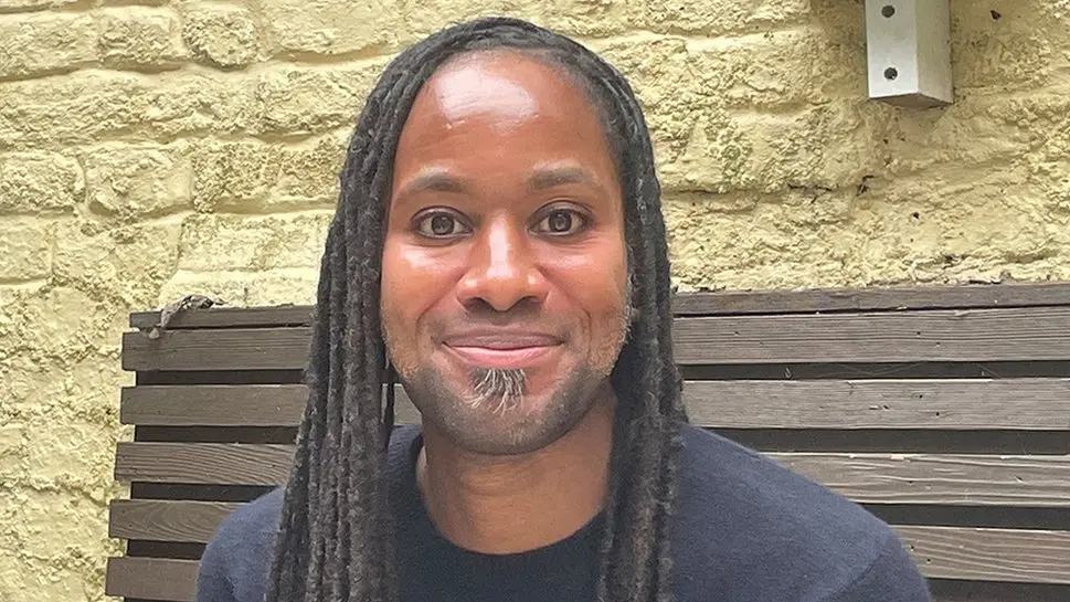 A man with dreadlocks smiles at the camera as he sits on a wooden bench