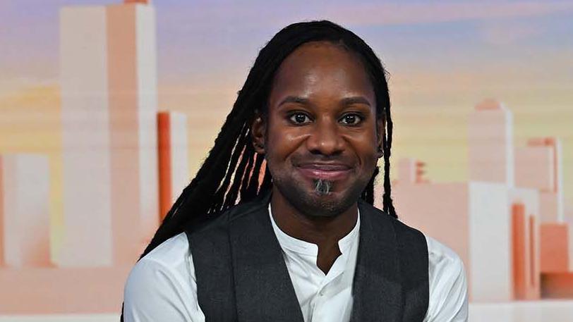 Head shot showing Professor Jason Arday sitting in the Sunday with Laura Kuennsberg studio. He is smiling at the camera with long black locs, wearing a white shirt with a grey waist coat.