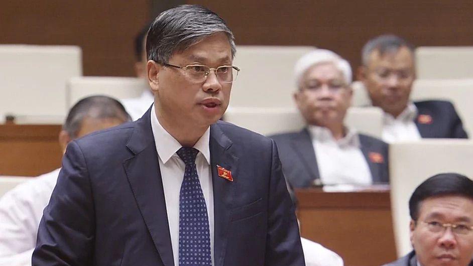 A man in a suit, tie and glasses, with a small red badge pinned to his lapel, stands inside what appears to be a parliament building