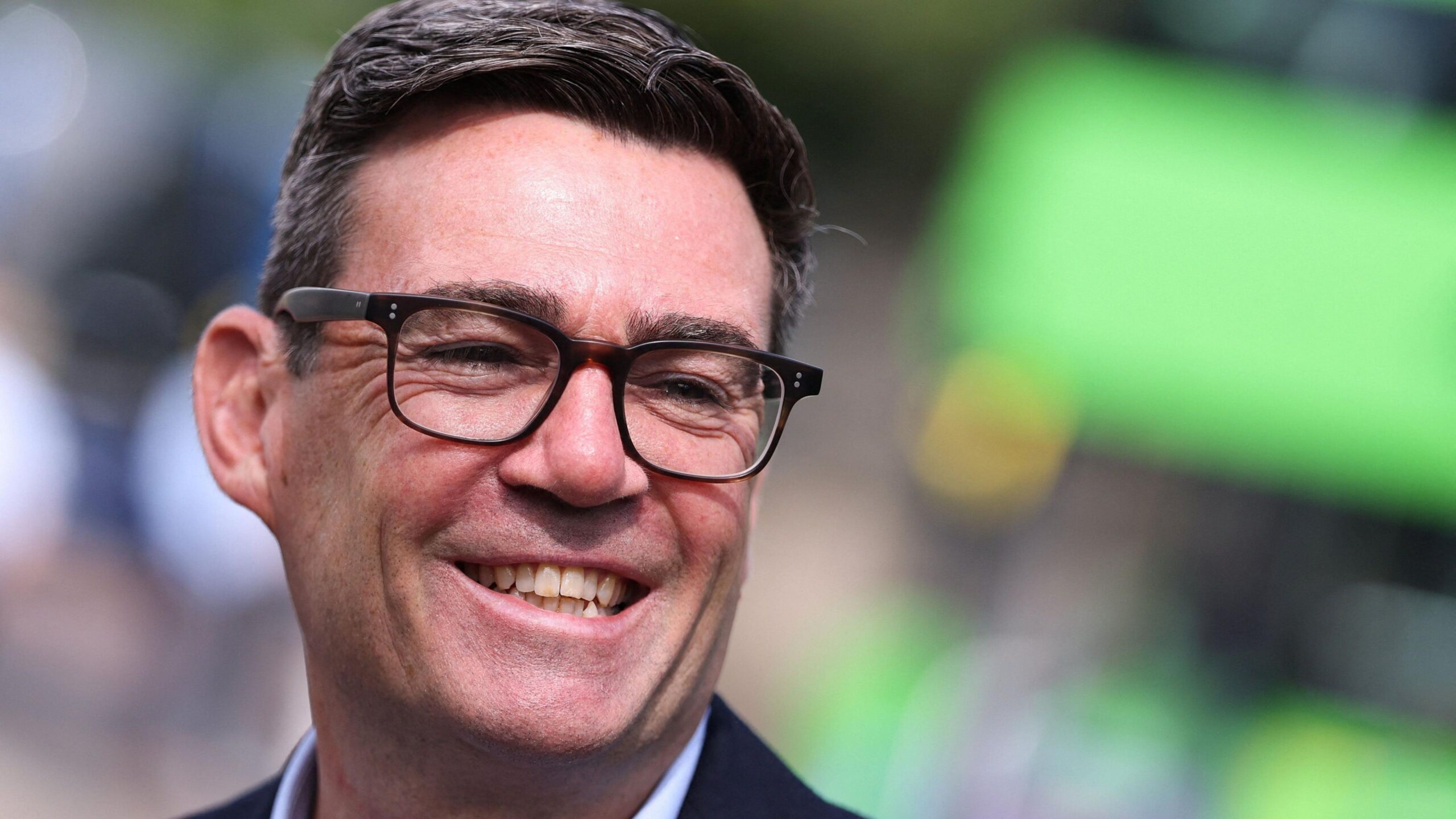 Andy Burnham is smiling and looking to the side. He has dark hair, brushed to one side and dark glasses.