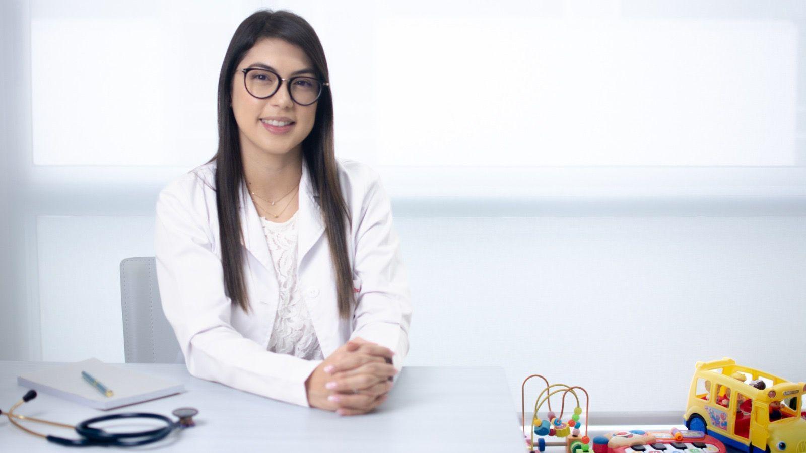 A woman with long brown hair wears a white medical coat and dark-framed glasses. She is seated indoors against a bright, predominantly white background that resembles a clinical setting. The image is a medium close-up portrait with soft lighting; no visible text, medical equipment, or other notable objects appear in the frame.