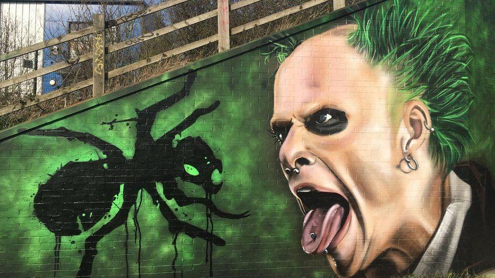 A mural of singer Keith Flint is painted on a green background on a large wall. The singer has his pierced tongue out and there is a large black ant near him.