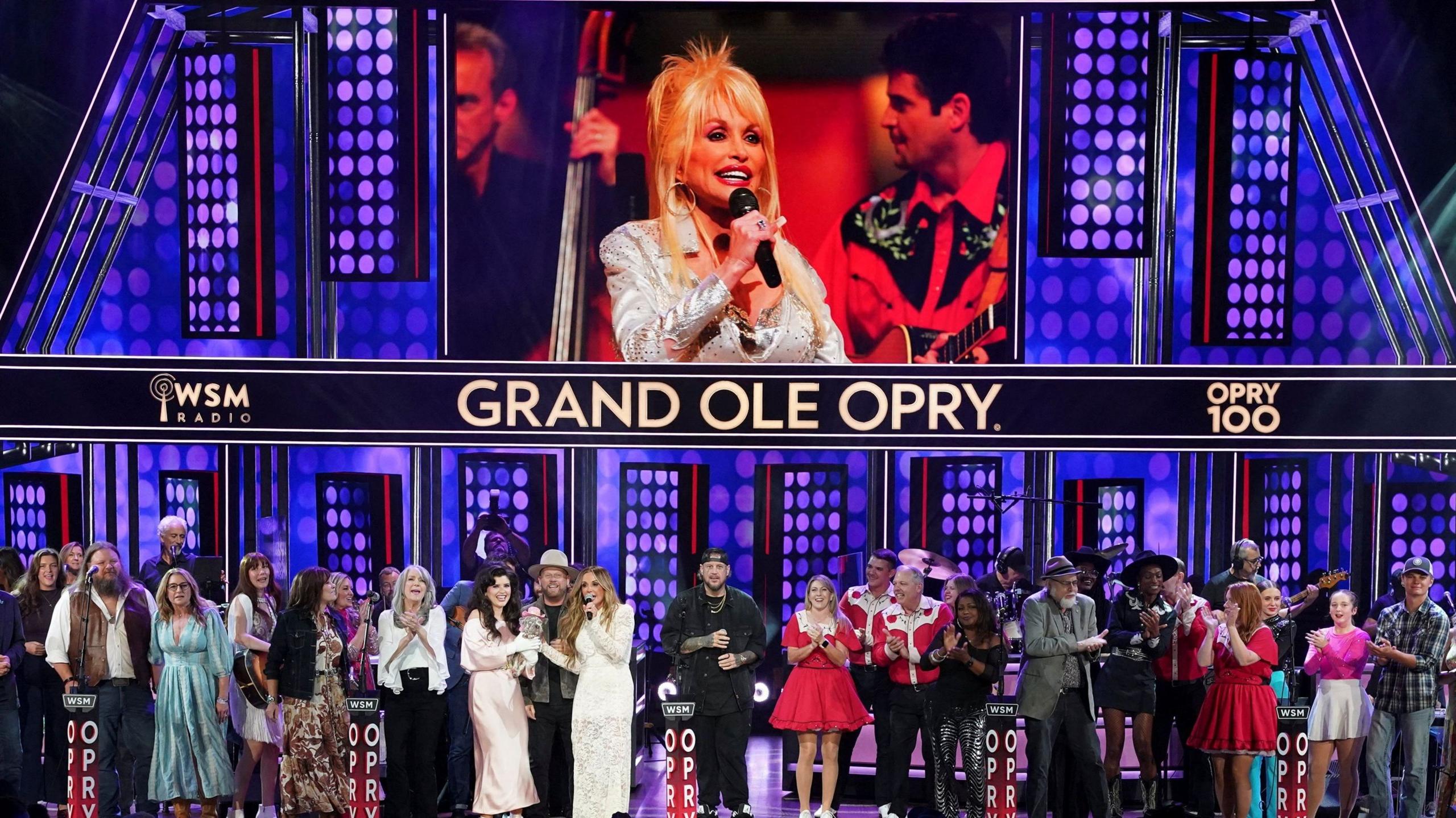 More than two dozen singers and musicians in a row on stage below a large screen showing a photo of Dolly Parton and the Grand Ole Opry name