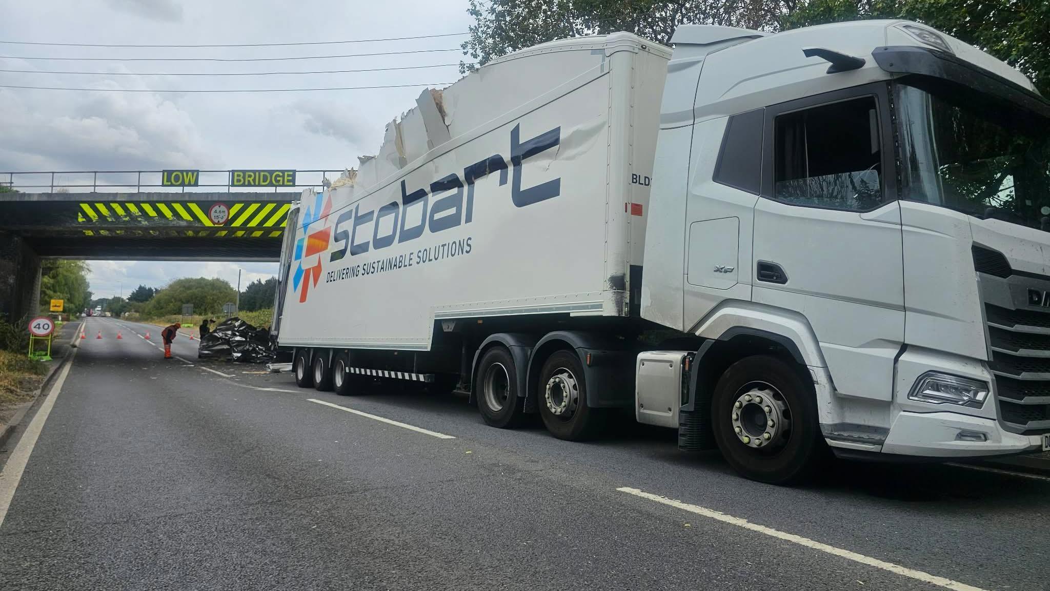 A road under a railway bridge is in the distance and at the front of the photograph is a lorry on the road with a damaged roof. There is debris and cones under the bridge.