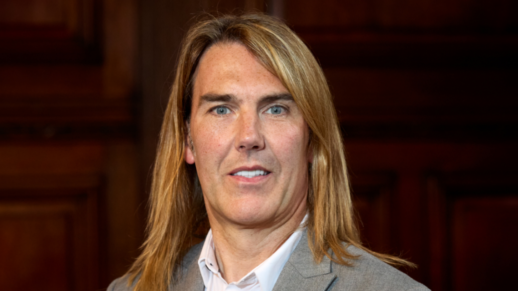 A man with long golden hair looking into the camera wearing a grey suit and white shirt.