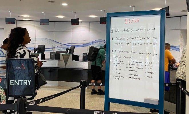 A hand-written whiteboard details the cancellations to trains, in the customer ticket office. On the right there is a sign that says Entry, behind which passengers are waiting to go to the counter.