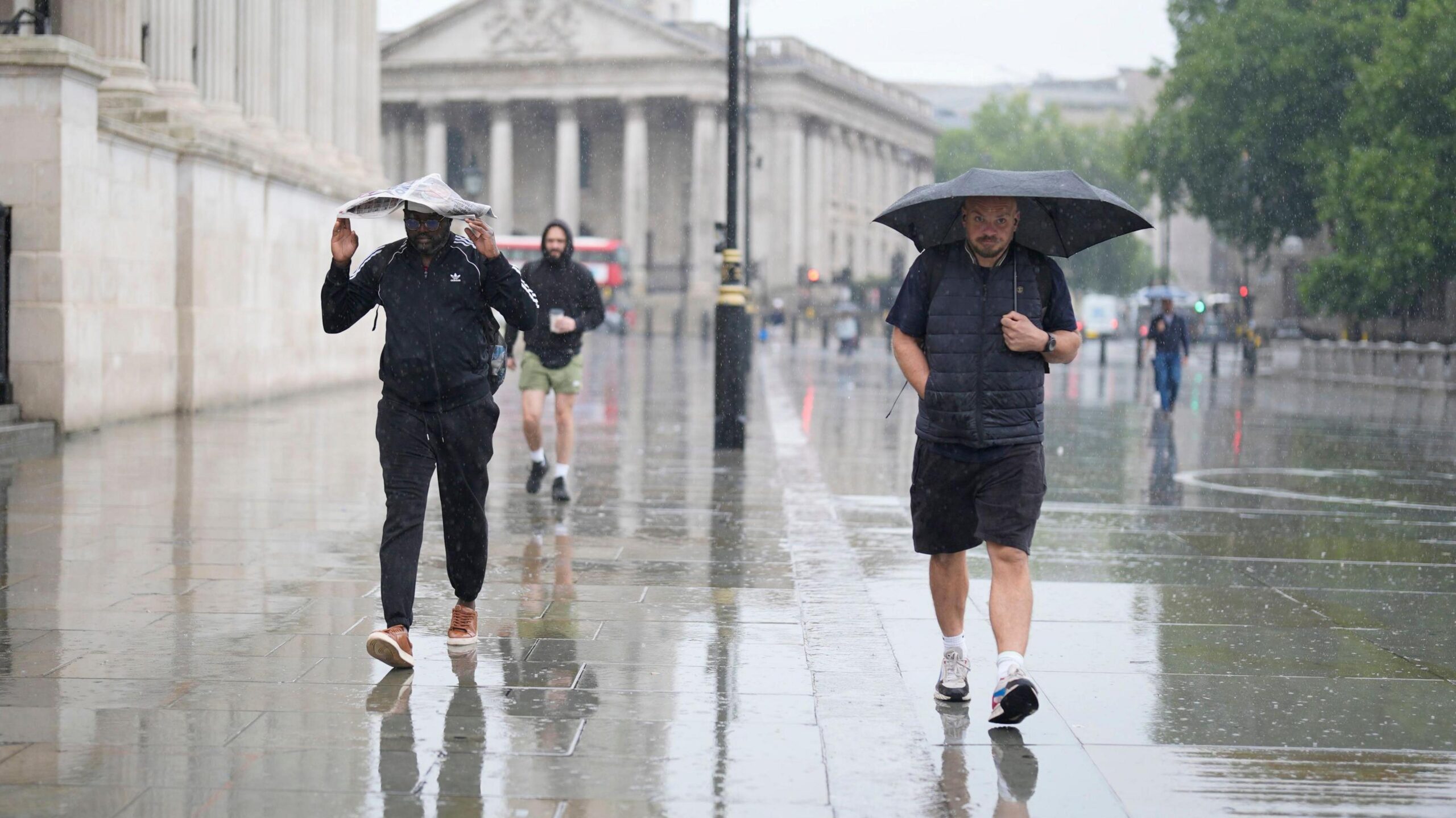 A man holding an umbrella and another man sheltering under a newspaper, walking down a street in the rain, with a city scene in the background