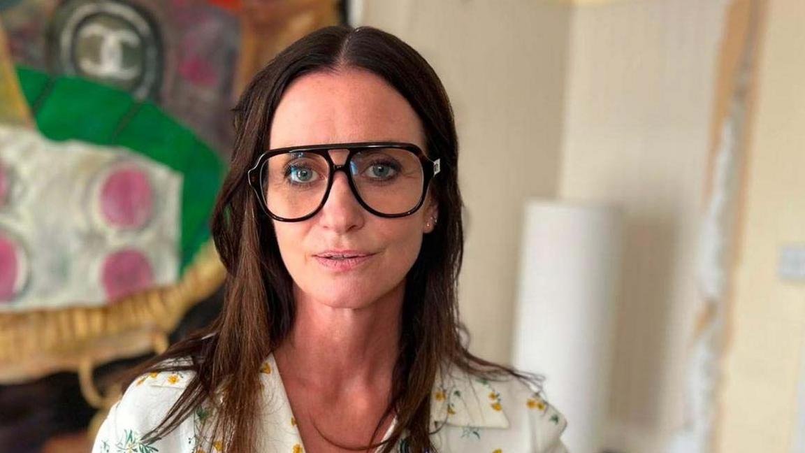 A woman with long dark hair looks at the camera. She is wearing large black glasses