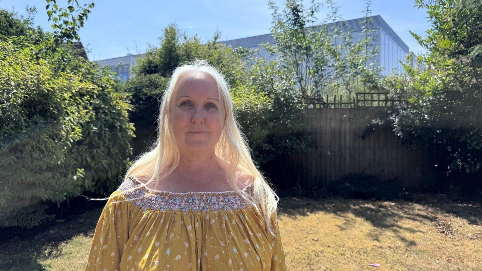 A woman with long blonde hair stands in her garden. Behind her, beyond her garden fence, is a large data centre which looks like a tall warehouse