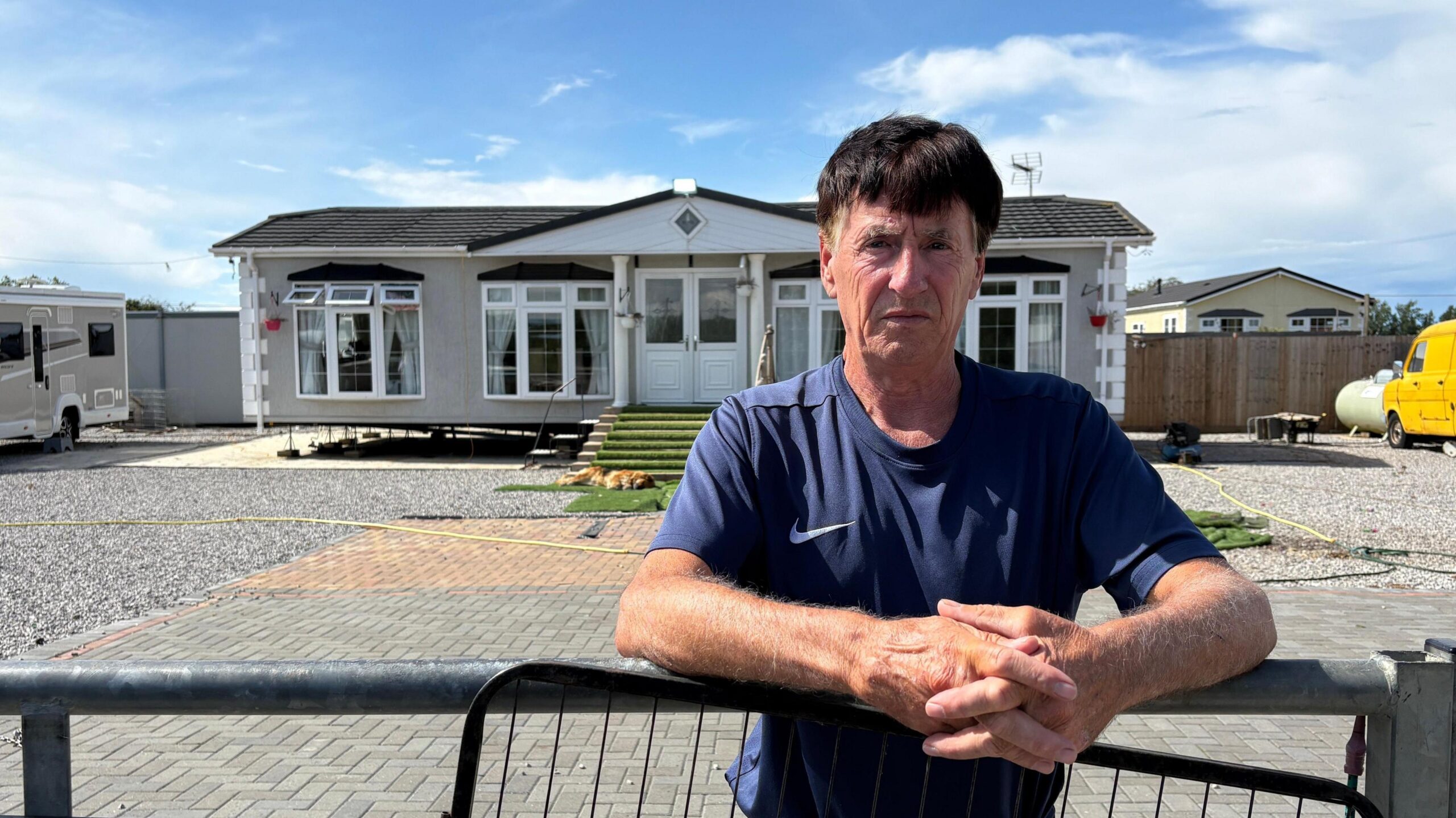 Clifford Codona leaning on a gate and looking direct to camera. He is standing outside, with a static caravan in the background. While the caravan is centred, Codona is positioned just to the right of centre. His forearms are resting on the top of the gate and his hands are clasped together in front of him. He is wearing a blue T-shirt. He has dark hair with greying temples.